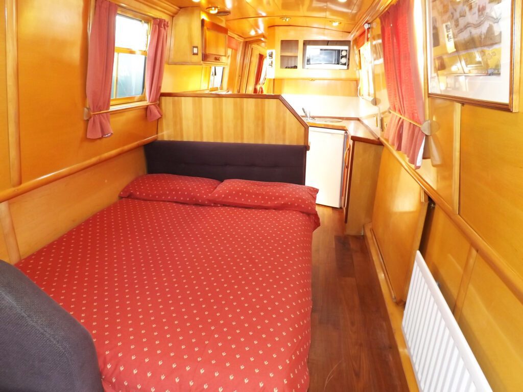 Double bed and galley kitchen inside a narrowboat with red bedding and wooden finish.