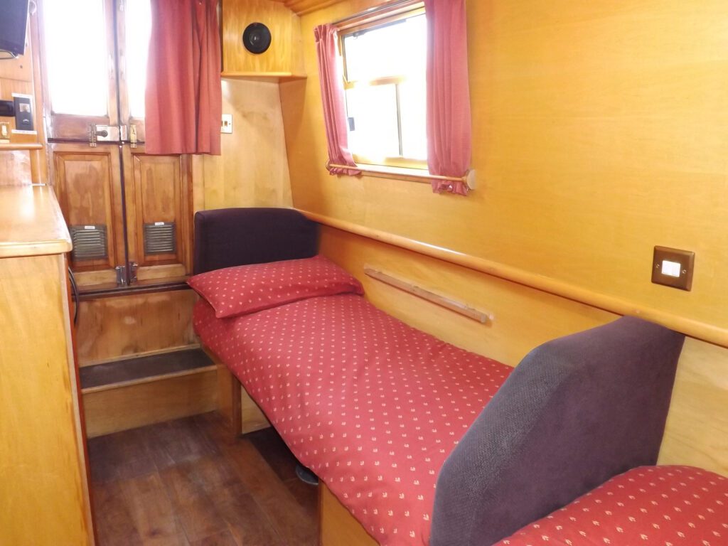 Single bed cabin inside a narrowboat with red bedding, wooden walls, and window with curtains.